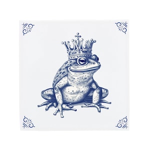 May include: A blue and white ceramic tile with a detailed illustration of a frog wearing a gold crown. The frog is sitting on a white background with blue decorative accents.