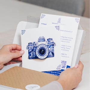 Peut inclure: Une cam&eacute;ra blanche avec des motifs floraux bleus et blancs, ressemblant &agrave; de la fa&iuml;ence de Delft, est pr&eacute;sent&eacute;e dans une image en gros plan. La cam&eacute;ra est sur une carte blanche avec le texte "The Dutch Project" et un code QR.