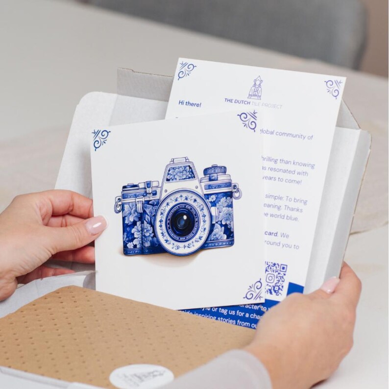 Peut inclure: Une cam&eacute;ra blanche avec des motifs floraux bleus et blancs, ressemblant &agrave; de la fa&iuml;ence de Delft, est pr&eacute;sent&eacute;e dans une image en gros plan. La cam&eacute;ra est sur une carte blanche avec le texte "The Dutch Project" et un code QR.