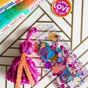 May include: A clear, rectangular pouch filled with colorful sequins, small paper cutouts, and a pink tassel with orange polka dot ribbon. The pouch is next to books with the text "Katherine Center" and a round sticker that says "Love is good for you."