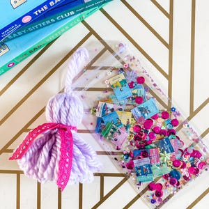 May include: A handmade lavender yarn tassel with a pink lace accent next to a clear pouch filled with miniature book cutouts, sequins, and confetti. The Baby-Sitters Club books are stacked in the background.