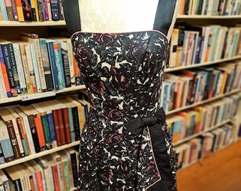 Op de afbeelding: Een zwart-witte bloemenjurk met een roze bies en een zwarte strik in de taille. De jurk is op een mannequin voor een boekenkast.