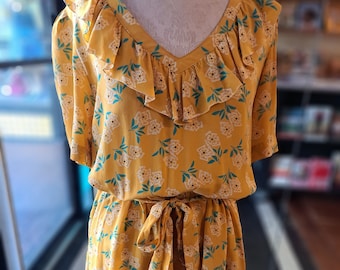 Op de afbeelding: Een gele bloemenjurk met een gerimpelde halslijn en een strik in de taille.