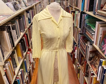 Op de afbeelding: Een vintage lichtgele jurk met een kraag, knoopsluiting en doorschijnende mouwen. De jurk heeft een kanten lijfje en een zwierige rok met een delicate afwerking. De jurk wordt tentoongesteld op een mannequin in een boekhandel.