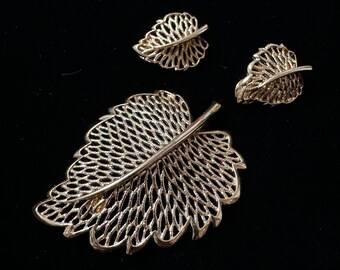 Op de afbeelding: Een gouden broche in de vorm van een blad en bijpassende oorbellen. De broche is groot en heeft een delicaat, ingewikkeld ontwerp. De oorbellen zijn kleiner en hebben een vergelijkbaar ontwerp.