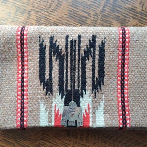 May include: A rectangular woven clutch in tan, black, red, and white. The central design features a black geometric pattern, flanked by red and white striped borders. A silver-colored metal emblem of a bird is attached to the front.