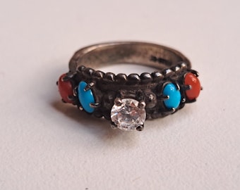 Op de afbeelding: Een zilverkleurige ring met een centrale heldere steen en afwisselend rode en turkooizen stenen. De ring heeft een getextureerde band en decoratieve zettingen, wat een vintage of Southwestern stijl creëert.