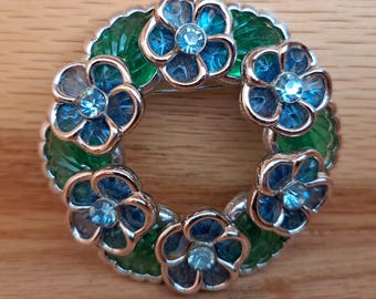 Op de afbeelding: Een zilverkleurige broche in de vorm van een krans met zes blauwe en groene bloemen. Elke bloem heeft een helder centrum met een blauwe strasssteen.