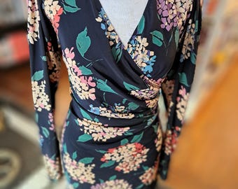 Op de afbeelding: Een marineblauwe wikkeljurk met lange mouwen en een bloemenpatroon. De jurk heeft een V-hals en is versierd met roze, witte en paarse bloemen en groene bladeren. De jurk wordt gepresenteerd op een mannequin.
