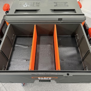 May include: A gray Klein Tools Modbox storage unit with an open drawer featuring two bright orange vertical dividers. The interior base is black with a molded logo. The exterior has orange side latches and the Klein Tools Modbox logo on the front.