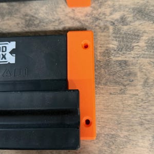 May include: Close-up of two black and orange woodworking tools. The black components have a rectangular shape with grooves, and the orange pieces are rectangular with two holes. The tools are on a wooden surface.