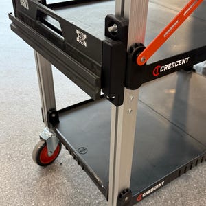 May include: A mobile utility cart featuring silver aluminum vertical supports and black plastic shelves. The cart includes a black Klein Tools Modbox attachment, a bright orange support arm, and black wheels with red centers. The Crescent brand logo is visible.