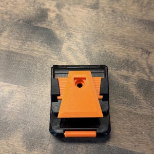 May include: A black and orange tool with a triangular orange piece in the center. The tool has a rectangular shape with a small orange handle at the bottom. The tool is sitting on a wooden surface.