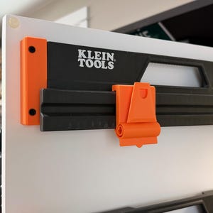 May include: A black and orange Klein Tools wall-mounted storage rail system featuring a horizontal track with a sliding orange clip. The black rail displays the white Klein Tools logo and is secured to a white panel with orange end caps.