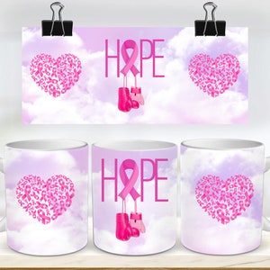 May include: White ceramic mug with a pink ribbon design. The mug features a heart made of pink ribbons, the word "HOPE" with a pink ribbon hanging from it, and pink boxing gloves. The background is a light purple and white cloudy sky.