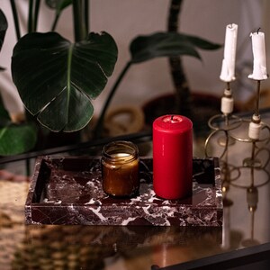 May include: A red candle and a brown glass jar candle are on a dark red marble tray. The tray is on a glass table with a gold metal candle holder with two white candles.