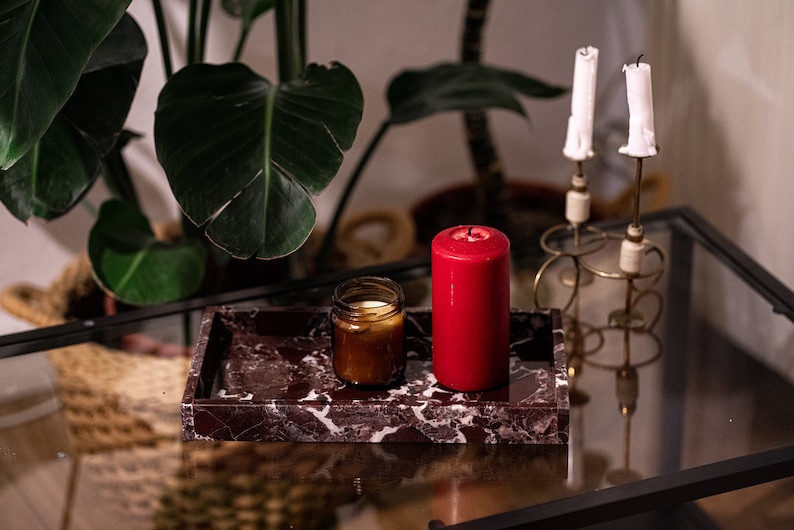 May include: A red candle and a brown glass jar candle are on a dark red marble tray. The tray is on a glass table with a gold metal candle holder with two white candles.