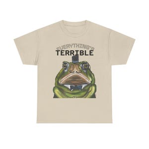 Peut inclure: T-shirt beige avec une illustration d'une grenouille verte portant un chapeau haut de forme et un nœud papillon. Le texte « EVERYTHING'S TERRIBLE » est imprimé au-dessus de la tête de la grenouille. Le design est centré sur le t-shirt.