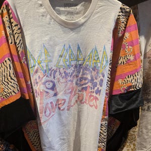 May include: Cream-colored Def Leppard graphic tee with the words "Love Bites" in pink. The sleeves feature a tiger stripe and floral pattern in orange, pink, and black. The shirt is hanging on a black hanger.