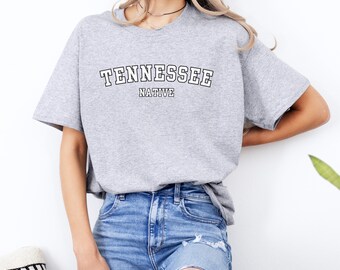 May include: A gray t-shirt with the text "Tennessee Native" printed in white on the front. The shirt is being worn by a person with long blonde hair and blue jeans.