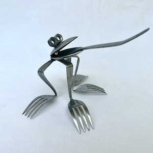 Puede incluir: Una escultura de rana de plata hecha con cubiertos reciclados. La rana tiene una lengua larga y está sentada sobre una superficie blanca.