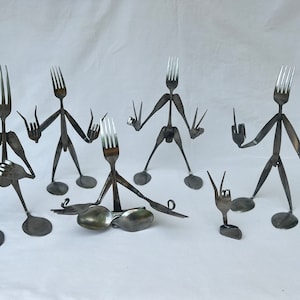 Op de afbeelding: Een groep van zes grillige metalen sculpturen gemaakt van vorken en lepels. De figuren hebben vorkkoppen, lepelvoeten en armen en benen van de handvatten van de gebruiksvoorwerpen. Elke figuur poseert in een ander gebaar.