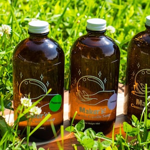 May include: Three brown glass bottles of Maker's! Cosmic Castile Soap, with white lids and labels. The bottles are on a wooden surface and surrounded by green grass. The labels feature a hand holding a sphere with stars around it. The scents are Peppermint, Lavender, and Orange Citrus.