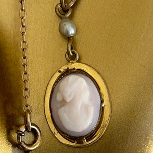 May include: A gold chain necklace with a cameo pendant featuring a white cameo profile portrait. The pendant is surrounded by a gold frame and has a single pearl bead above it.