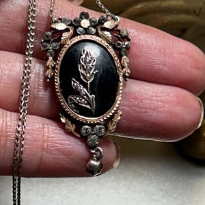 May include: A silver chain necklace with a black cameo pendant featuring a floral design. The pendant is surrounded by a silver frame with a rose gold trim and small clear stones.