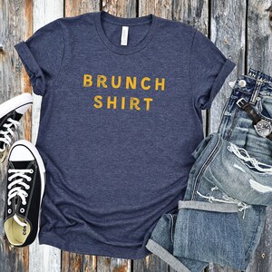 K&ouml;nnte beinhalten: Ein marineblaues T-Shirt mit dem Text "BRUNCH SHIRT" in Gelb.
