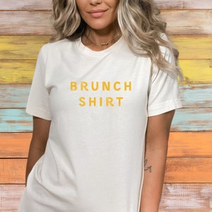 K&ouml;nnte beinhalten: Ein wei&szlig;es T-Shirt mit dem Aufdruck "BRUNCH SHIRT" in Gelb.