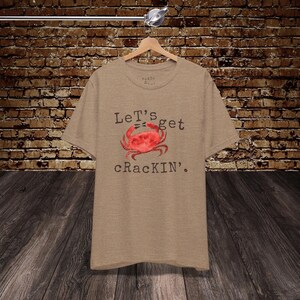 May include: A beige t-shirt featuring the phrase "Let's get crackin'" and a red crab illustration. The t-shirt is displayed on a wooden hanger. The background is a brick wall and wooden floor. The shirt is made of a soft material.