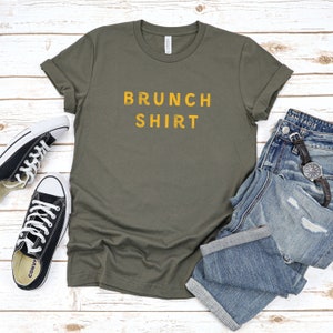 K&ouml;nnte beinhalten: Ein gr&uuml;nes T-Shirt mit dem Text "BRUNCH SHIRT" in gelben Buchstaben. Das Shirt liegt flach auf einer wei&szlig;en Holzoberfl&auml;che mit einem Paar schwarzer Turnschuhe und einer blauen Jeans.