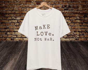Könnte beinhalten: Weißes T-Shirt mit der Aufschrift "MaKe LOVE. Not WaR." in braunen Buchstaben. Das Shirt hängt an einem Holzbügel vor einem Hintergrund aus Backsteinmauerwerk.