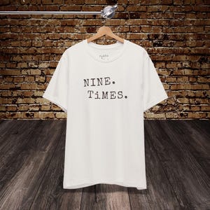 Peut inclure: T-shirt blanc avec le texte "NINE. TIMES." en noir, présenté sur un cintre en bois. Le t-shirt est suspendu devant un mur de briques et un sol en bois. Le t-shirt a un col rond et des manches courtes.