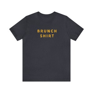 K&ouml;nnte beinhalten: Ein dunkelgraues T-Shirt mit dem Aufdruck "BRUNCH SHIRT" in Gelb.