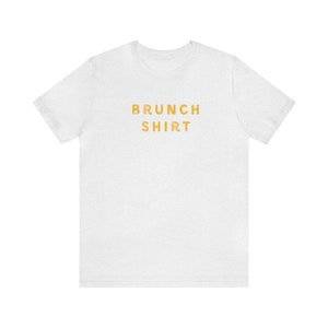 K&ouml;nnte beinhalten: Wei&szlig;es T-Shirt mit dem Aufdruck "BRUNCH SHIRT" in Orange.