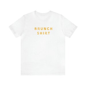 K&ouml;nnte beinhalten: Wei&szlig;es T-Shirt mit dem Aufdruck "BRUNCH SHIRT" in Orange.