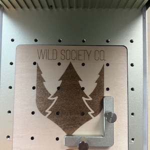 Op de afbeelding: Een close-up van een houten bord met een lasergesneden ontwerp van een dennenboom en de tekst "WILD SOCIETY CO.". Het bord is bevestigd aan een metalen frame met een zilverkleurige verstelbare klem.
