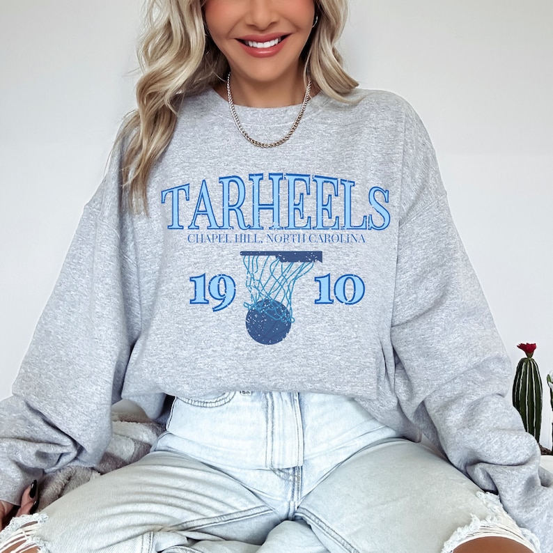 K&ouml;nnte beinhalten: Grauer Pullover mit einer blauen Grafik eines Basketballkorbs und dem Text "TARHEELS CHAPEL HILL, NORTH CAROLINA 19 10".