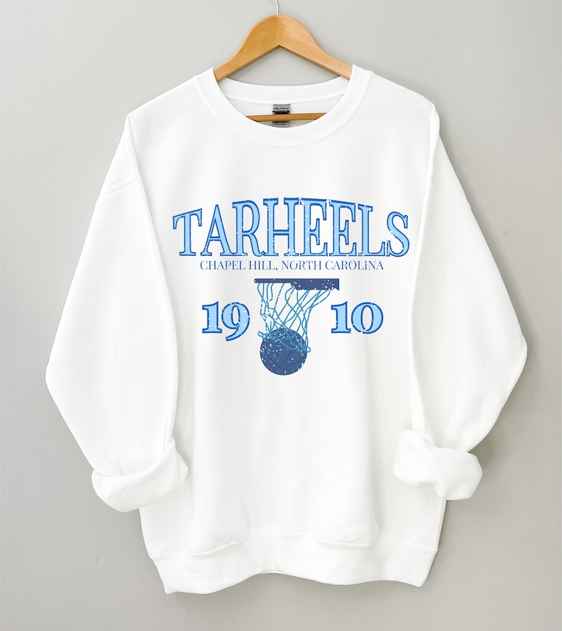 K&ouml;nnte beinhalten: Wei&szlig;er Pullover mit einem blauen Grafikdruck eines Basketballs, der durch einen Korb geht. Der Text "Tar Heels" befindet sich &uuml;ber der Grafik, und "Chapel Hill, North Carolina" befindet sich unter der Grafik. Die Zahlen "19" und "10" befinden sich auf beiden Seiten der Grafik.