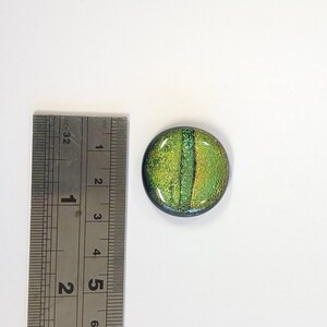 Könnte beinhalten: Eine runde, grüne und goldene Glasfliese mit einer strukturierten Oberfläche. Die Fliese hat einen Durchmesser von etwa 2,5 cm.