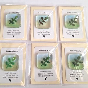 Puede incluir: Seis amuletos de bolsillo, cada uno con un diseño de vidrio cuadrado con una planta verde sobre un fondo azul y verde. Cada amuleto está empaquetado en una tarjeta color crema con el texto "Pocket Charm" y un mensaje.