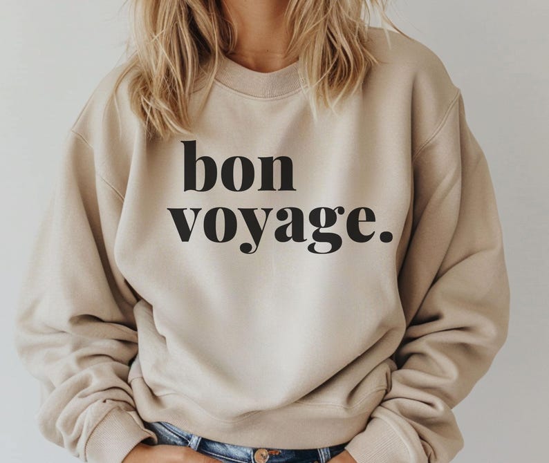 May include: A beige crew neck sweatshirt with the words "bon voyage" printed in black. The text is in a bold, sans-serif font. The long-sleeved top is a neutral colour.