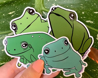May include: Three green frog stickers with white outlines. The frogs are in different poses, with one looking up, one looking straight ahead, and one looking down. The stickers are on a green leaf background.