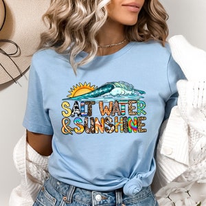May include: A light blue t-shirt with a colorful graphic of a sun, wave, and the text "Salt Water & Sunshine".