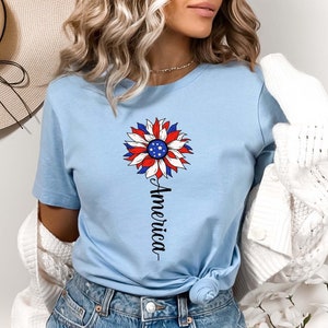Puede incluir: Una camiseta azul claro con un dise&ntilde;o de girasol rojo, blanco y azul. El girasol tiene una peque&ntilde;a bandera estadounidense en el centro. La palabra "America" est&aacute; escrita verticalmente debajo del girasol.