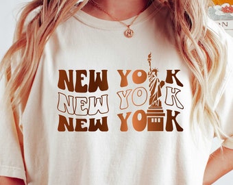 May include: A beige t-shirt with a graphic print of the Statue of Liberty and the text "NEW YORK" repeated three times in a retro font.
