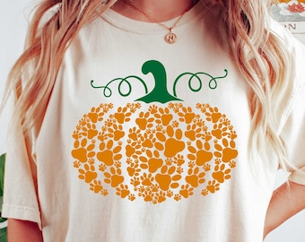 Könnte beinhalten: Ein weißes T-Shirt mit einem Kürbisdesign aus Pfotenabdrücken. Der Kürbis ist orange mit einem grünen Stiel und einer Ranken. Das Shirt ist überdimensioniert und hat eine lockere Passform.