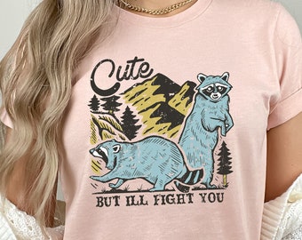 May include: A pink t-shirt with a graphic of two raccoons in a forest setting. The text on the shirt reads "Cute but I'll fight you."